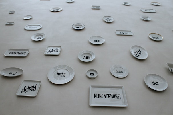 A collection of white ceramic plates and rectangular dishes displayed on a wall. Each piece has a German word or phrase printed in bold, black font. The words appear in various sizes and orientations, adding an abstract and artistic element to the display.