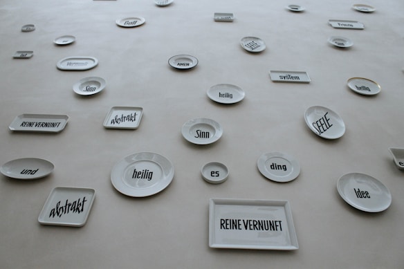 A collection of white ceramic plates and rectangular dishes displayed on a wall. Each piece has a German word or phrase printed in bold, black font. The words appear in various sizes and orientations, adding an abstract and artistic element to the display.