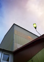 A corrugated metal building is viewed from an upward angle, featuring a section of the roofline against a sky transitioning from blue to pale pink. A single street lamp is visible, with a greenish tint to its light. The building shows various shades of muted yellow and green panels.