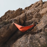 An orange hammock is suspended between rugged rock formations. The hammock is attached securely to ropes on the cliff face. The rocky landscape suggests an outdoor, adventurous setting.