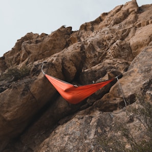 An orange hammock is suspended between rugged rock formations. The hammock is attached securely to ropes on the cliff face. The rocky landscape suggests an outdoor, adventurous setting.