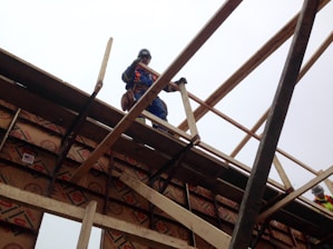 A construction worker wearing a safety harness stands on a wooden framework at an elevated height. The structure is made of wooden beams and boards, with safety equipment visible around the worker. Part of another worker can be seen in the background, wearing a hard hat and safety vest.