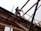 A construction worker wearing a safety harness stands on a wooden framework at an elevated height. The structure is made of wooden beams and boards, with safety equipment visible around the worker. Part of another worker can be seen in the background, wearing a hard hat and safety vest.