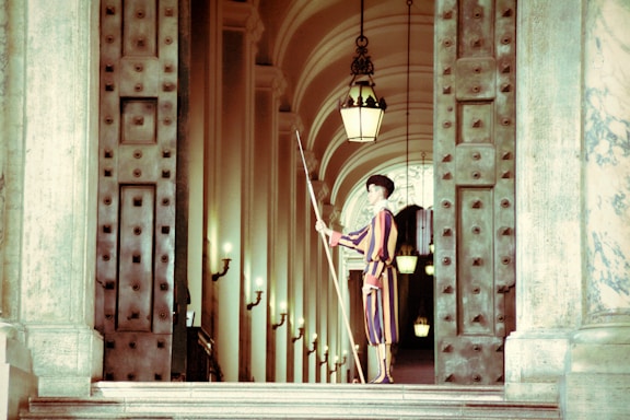 A guard dressed in a colorful uniform stands at attention in a grand corridor. The corridor features large, ornate metal doors and multiple hanging lanterns. The arched ceiling and walls are adorned with decorative elements and lamps.