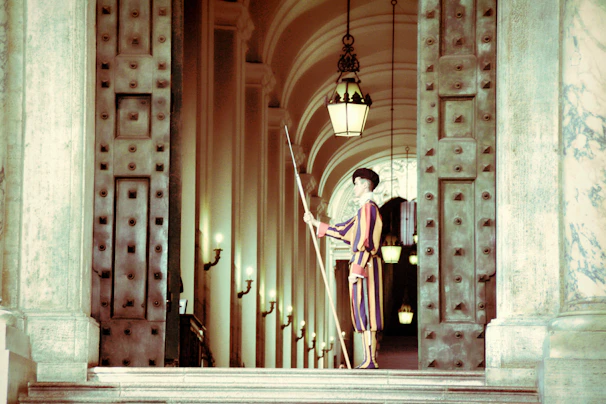 A guard dressed in a colorful uniform stands at attention in a grand corridor. The corridor features large, ornate metal doors and multiple hanging lanterns. The arched ceiling and walls are adorned with decorative elements and lamps.