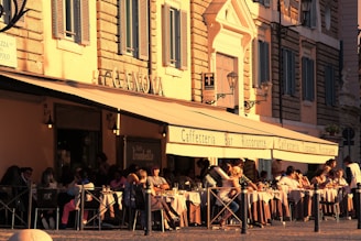 A bustling outdoor cafe with people seated at tables covered with white tablecloths. The building features large awnings with signage for a cafe and restaurant. The architecture includes stone walls and decorative window shutters, with the warm golden light of the setting sun casting a cozy atmosphere.
