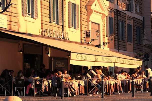A bustling outdoor cafe with people seated at tables covered with white tablecloths. The building features large awnings with signage for a cafe and restaurant. The architecture includes stone walls and decorative window shutters, with the warm golden light of the setting sun casting a cozy atmosphere.