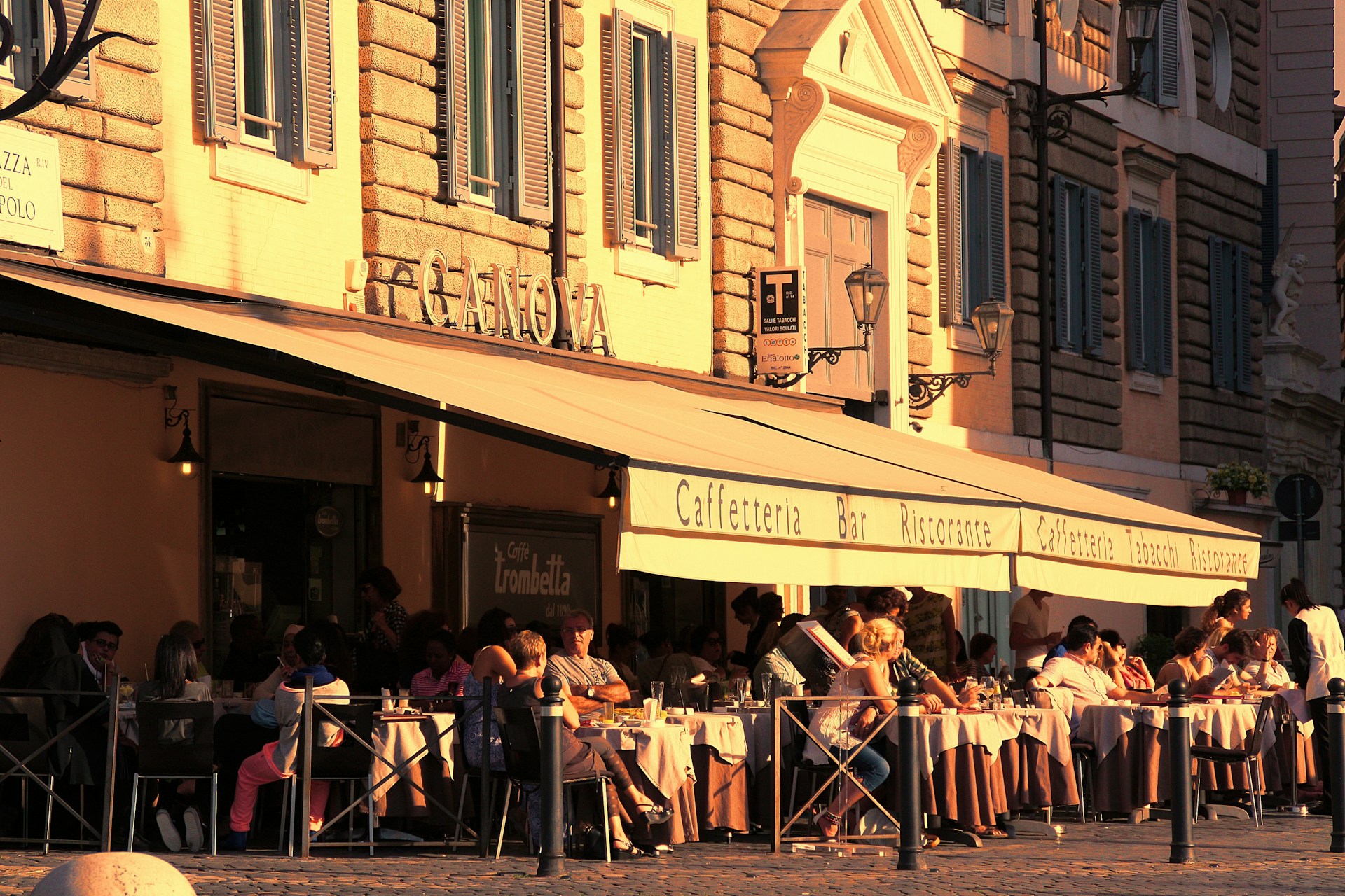 A bustling outdoor cafe with people seated at tables covered with white tablecloths. The building features large awnings with signage for a cafe and restaurant. The architecture includes stone walls and decorative window shutters, with the warm golden light of the setting sun casting a cozy atmosphere.