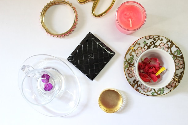 A variety of decorative items are arranged on a white surface, including a gold bracelet with pink accents, a pink candle, a black square box with a textured surface, a clear glass teacup with a saucer containing purple petals, and a porcelain teacup with a floral design holding red petals. There is also a golden ring with a reflective center.