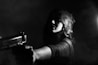 A black and white image shows a woman with curled hair, wearing a hat, and holding a gun aimed directly at the camera. The scene is dimly lit, casting dramatic shadows across her face, creating a noir effect.