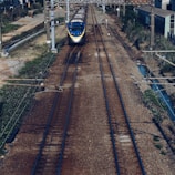 A passenger train approaches on a set of parallel tracks with overhead power lines visible. The surrounding area features grassy and shrub-lined paths, with utility poles and a mix of industrial and rural landscapes in the background.