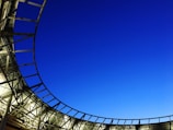 A modern architectural structure with a curved metal framework partially enclosing a section of vibrant blue sky. The design features a combination of glass and metal, creating a sleek and open appearance.