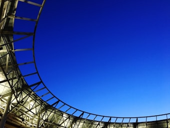 A modern architectural structure with a curved metal framework partially enclosing a section of vibrant blue sky. The design features a combination of glass and metal, creating a sleek and open appearance.