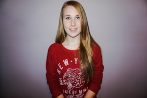 A person with long blond hair is wearing a red sweatshirt with white text and a graphic image of a panther head. The background is a simple, neutral-colored wall.