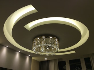 A contemporary ceiling design featuring illuminated curved lines that form a circular pattern around a luxurious chandelier. The chandelier is adorned with numerous crystal ornaments that elegantly reflect light, creating a sophisticated ambiance. The room is softly lit with recessed lighting, contributing to the modern aesthetic.