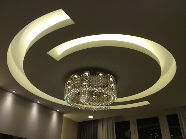 A contemporary ceiling design featuring illuminated curved lines that form a circular pattern around a luxurious chandelier. The chandelier is adorned with numerous crystal ornaments that elegantly reflect light, creating a sophisticated ambiance. The room is softly lit with recessed lighting, contributing to the modern aesthetic.