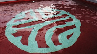 A swimming pool with clear, rippling water reflecting the light. At the bottom, a large, abstract design is visible, featuring a combination of green and aqua hues against a red-tiled background.