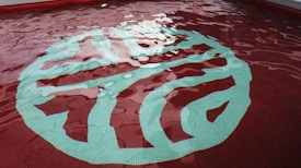 A swimming pool with clear, rippling water reflecting the light. At the bottom, a large, abstract design is visible, featuring a combination of green and aqua hues against a red-tiled background.