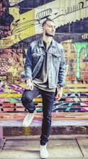 A person stands confidently against a colorful graffiti wall, wearing a denim jacket, dark jeans, and white sneakers. The background features vibrant and abstract street art with patterns and drips in various colors.