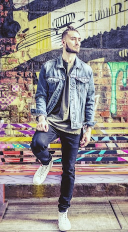 A person stands confidently against a colorful graffiti wall, wearing a denim jacket, dark jeans, and white sneakers. The background features vibrant and abstract street art with patterns and drips in various colors.