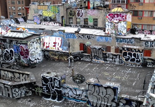 A rooftop densely covered in vibrant graffiti art with various styles and colors. The walls and surfaces are filled with tags, throw-ups, and large murals, displaying the urban art culture. The buildings are in an urban environment with visible windows and some air conditioning units.