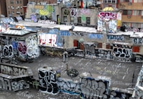 A rooftop densely covered in vibrant graffiti art with various styles and colors. The walls and surfaces are filled with tags, throw-ups, and large murals, displaying the urban art culture. The buildings are in an urban environment with visible windows and some air conditioning units.