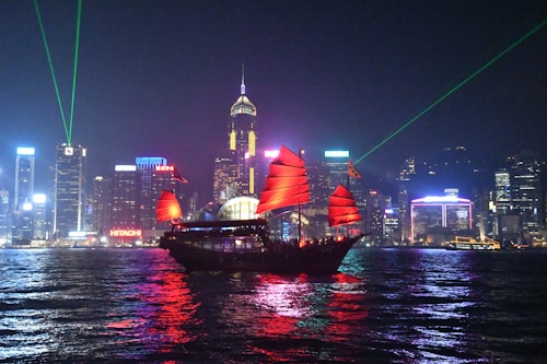 A traditional Chinese junk boat with bright red sails is sailing on a dark body of water against a backdrop of an illuminated city skyline. The city buildings feature colorful lights and green laser beams projecting into the sky, creating a vibrant and dynamic night scene.