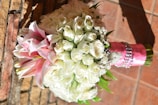 A bouquet of white roses mixed with pink lilies, wrapped in pink and silver ribbons, resting against a rustic stone wall. The arrangement is lush and carefully positioned, showcasing a harmonious blend of colors and textures.