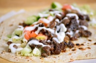 A close-up of a taco on a flour tortilla filled with seasoned meat, chopped lettuce, diced tomatoes, sliced cucumbers, and drizzled with white sauce. The tortilla has a slightly toasted appearance.