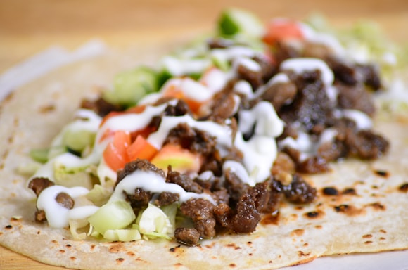 A close-up of a taco on a flour tortilla filled with seasoned meat, chopped lettuce, diced tomatoes, sliced cucumbers, and drizzled with white sauce. The tortilla has a slightly toasted appearance.