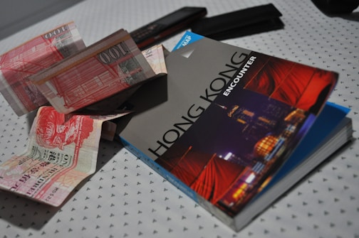 A Hong Kong travel guidebook is placed on a patterned surface alongside several Hong Kong banknotes. The guidebook features a vibrant cityscape on the cover, highlighting iconic red sails. The banknotes appear to be of different denominations and are casually scattered, suggesting themes of travel and exploration.