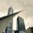 A striking architectural scene featuring modern skyscrapers with sleek glass exteriors. The design includes sharp angles and a prominent structure resembling wings or a futuristic canopy in the foreground. The sky is overcast, creating a dramatic contrast with the metallic sheen of the buildings.