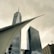 A striking architectural scene featuring modern skyscrapers with sleek glass exteriors. The design includes sharp angles and a prominent structure resembling wings or a futuristic canopy in the foreground. The sky is overcast, creating a dramatic contrast with the metallic sheen of the buildings.