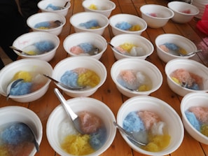 Several white bowls containing colorful shaved ice in shades of blue, pink, and yellow are arranged on a wooden table. Each bowl has a metal spoon resting inside, and the surface of the table is visible between the bowls. The setting appears casual and ready for serving.