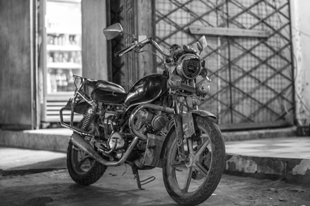 An old motorcycle parked indoors next to a refrigerator. The motorcycle has a rugged and worn appearance, with visible engine parts and a classic design. The background shows a large, closed gate with a crisscross pattern.