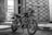 An old motorcycle parked indoors next to a refrigerator. The motorcycle has a rugged and worn appearance, with visible engine parts and a classic design. The background shows a large, closed gate with a crisscross pattern.