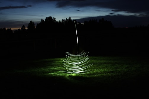 A spiral light structure hovers over a grassy field during twilight, creating an intriguing and futuristic ambiance with its smooth, glowing lines contrasting against the dark surroundings.