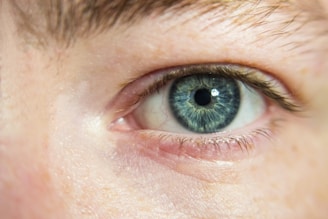A close-up of a blue human eye with detailed textures in the iris, surrounded by eyelashes and skin. Subtle light reflections are visible on the moist eye surface, enhancing the iridescent effect of the eye color.