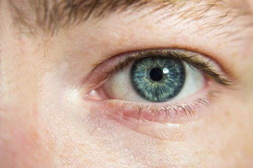 A close-up of a blue human eye with detailed textures in the iris, surrounded by eyelashes and skin. Subtle light reflections are visible on the moist eye surface, enhancing the iridescent effect of the eye color.