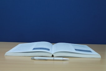 An open book is placed on a light wooden table with a pen lying in front of it. The background is a solid dark blue color, creating a simple and focused composition.