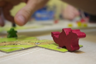 A close-up of a tabletop board game, featuring a red dragon-shaped game piece in focus on a square tile. In the background, a green meeple and another hand are slightly blurred, indicating gameplay. The surface is light-colored, and the scene conveys an intimate setting of game play.