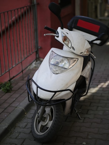 A white motor scooter is parked on a paved walkway, next to a red wall with metal railings. The scooter is equipped with a front grille for protection and a top box with a red accent, likely for carrying items. The lighting suggests it is outdoors, possibly in an urban setting.