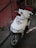 A white motor scooter is parked on a paved walkway, next to a red wall with metal railings. The scooter is equipped with a front grille for protection and a top box with a red accent, likely for carrying items. The lighting suggests it is outdoors, possibly in an urban setting.