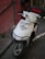 A white motor scooter is parked on a paved walkway, next to a red wall with metal railings. The scooter is equipped with a front grille for protection and a top box with a red accent, likely for carrying items. The lighting suggests it is outdoors, possibly in an urban setting.