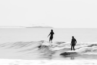 Two surfers are riding small waves in the ocean, both wearing wetsuits, and standing on their surfboards. The background is a serene open sea with a faint outline of a bridge or pier in the distance. The image uses monochrome tones, giving it a timeless and peaceful feel.