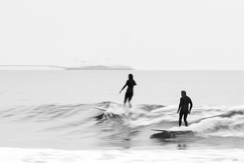 Two surfers are riding small waves in the ocean, both wearing wetsuits, and standing on their surfboards. The background is a serene open sea with a faint outline of a bridge or pier in the distance. The image uses monochrome tones, giving it a timeless and peaceful feel.