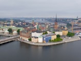 A picturesque urban landscape depicts a blend of historical and modern architecture situated on a waterfront. The scene features a variety of colorful buildings with spires and domes. The water reflects the structures, and several bridges connect the different areas. The atmosphere appears calm yet lively, with signs of both local activity and tourism.