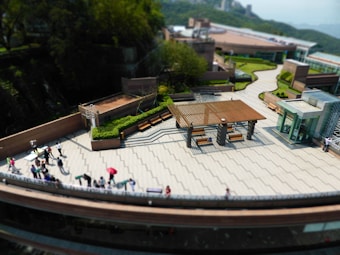 An elevated view of a rooftop terrace with a modern design. The area features pathways and seating arrangements, including benches under a wooden pergola. There are several people walking around, some using umbrellas for shade. The terrace is surrounded by greenery and buildings in the background.