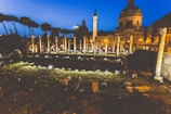 Ancient Roman ruins are illuminated at dusk, featuring a series of columns and stone structures in an archaeological site. The backdrop includes a prominent domed building with classical architecture. Soft lighting enhances the historic ambiance with a clear evening sky adding to the serene atmosphere.