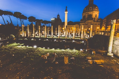 Ancient Roman ruins are illuminated at dusk, featuring a series of columns and stone structures in an archaeological site. The backdrop includes a prominent domed building with classical architecture. Soft lighting enhances the historic ambiance with a clear evening sky adding to the serene atmosphere.